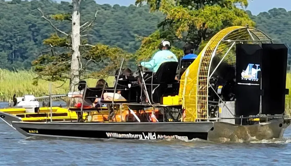 A group of people are enjoying a tour on an airboat labeled Williamsburg Wild Airboat Tours on a body of water surrounded by trees and greenery