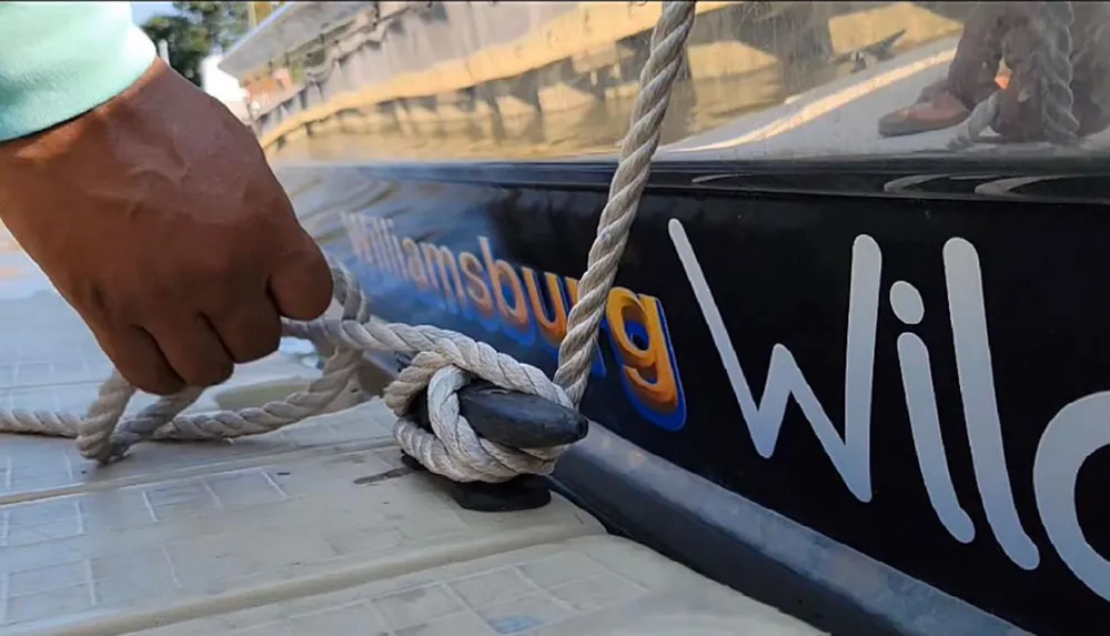 A person is tying a thick rope onto a cleat on a dock next to a boat with the partially visible name Williamsburg Wing