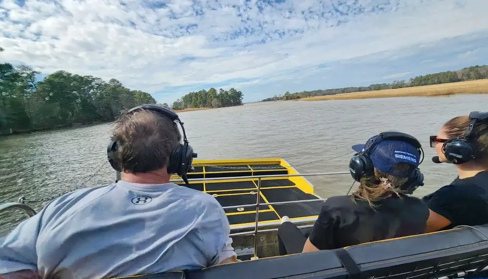 Three people wearing headphones are seated in a boat gliding on a river under a partly cloudy sky
