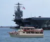 A tour boat named Miss Hampton II is cruising near a large naval aircraft carrier in a harbor