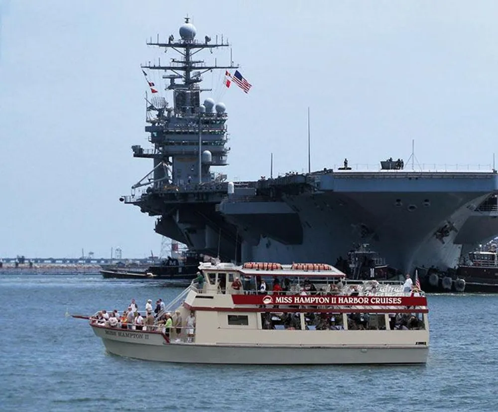 A tour boat named Miss Hampton II is cruising near a large naval aircraft carrier in a harbor