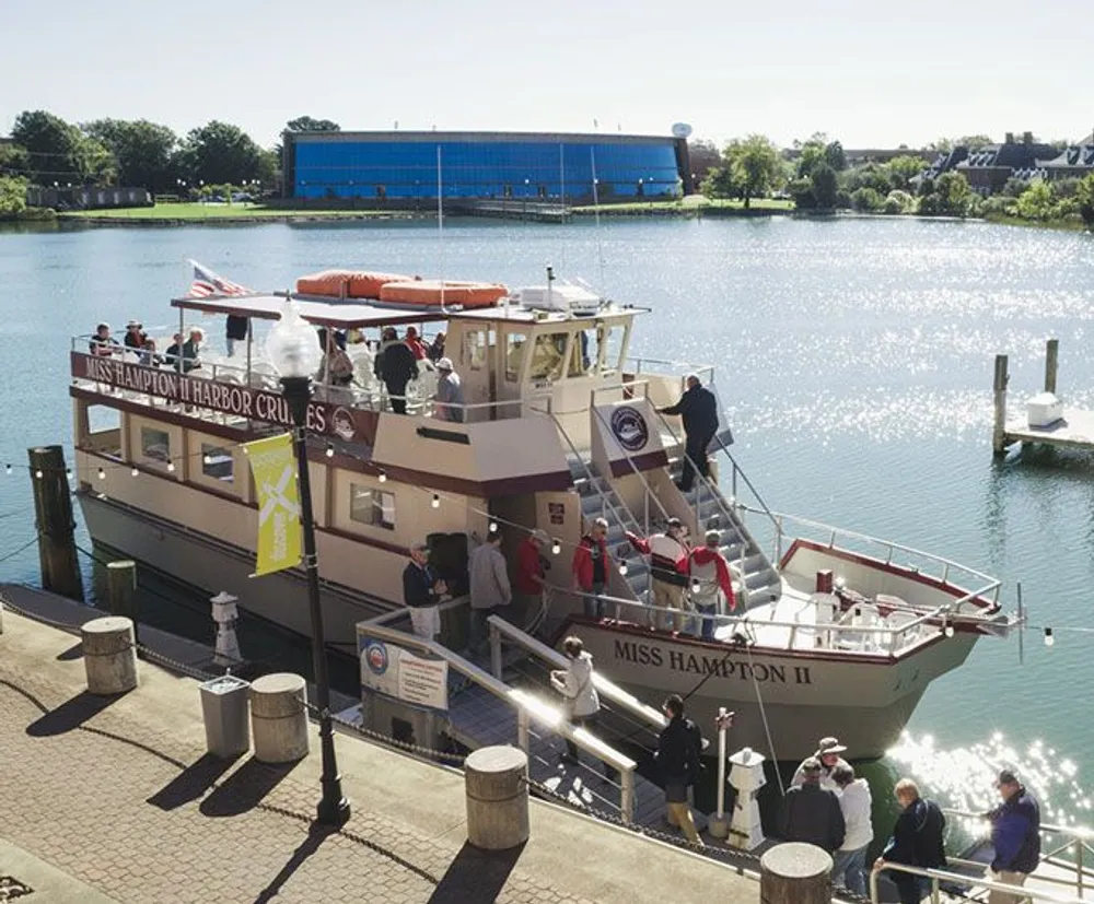 Passengers are boarding the Miss Hampton II tour boat on a sunny day at a calm harbor with a blue building in the background