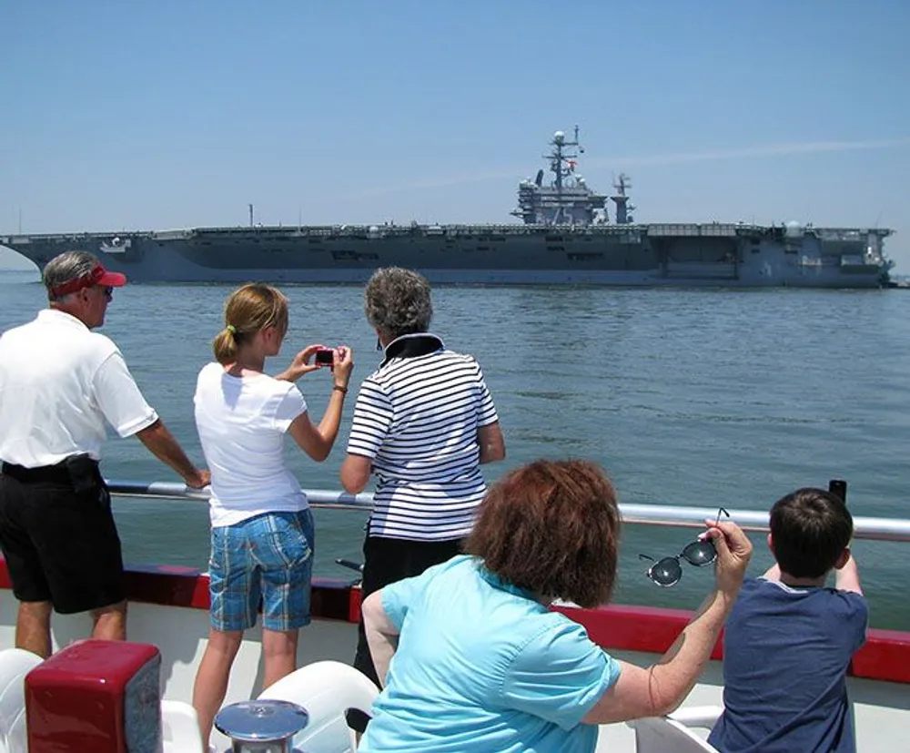 A group of tourists on a boat are observing and photographing a large naval aircraft carrier at sea