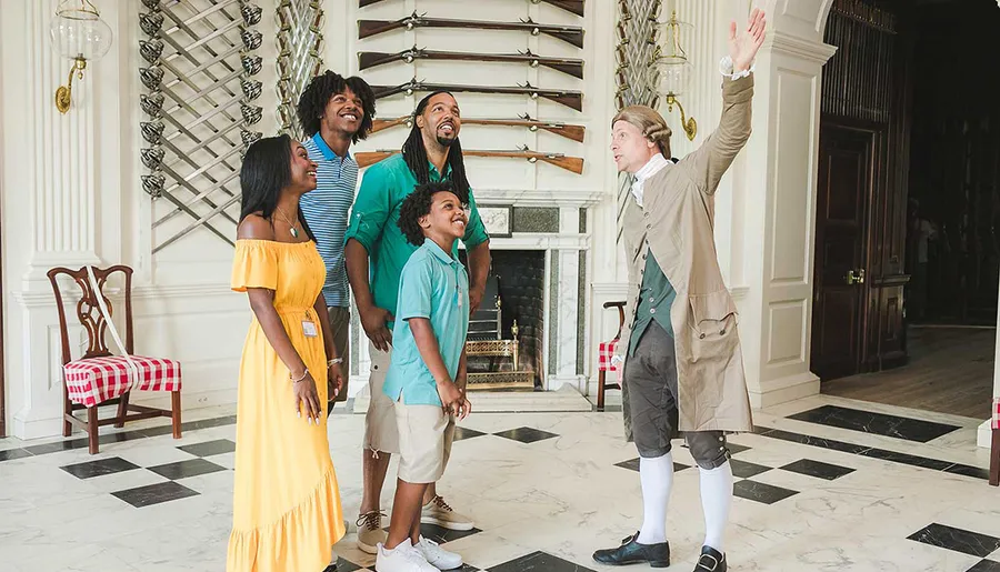 A family listens to a historical reenactor gesturing grandly while touring an elegant, colonial-style building.