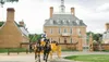 A horse-drawn carriage in historical attire is passing in front of an elegant colonial building with pedestrians nearby.