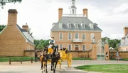 A horse-drawn carriage in historical attire is passing in front of an elegant colonial building with pedestrians nearby.