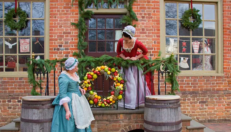 Two women in period costumes decorate a festive Christmas wreath on a brick building's balcony, while another woman watches from inside.