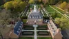 The image shows an aerial view of a large, symmetrical brick mansion with a neatly landscaped garden featuring geometric patterns and pathways.