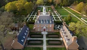 The image shows an aerial view of a large, symmetrical brick mansion with a neatly landscaped garden featuring geometric patterns and pathways.