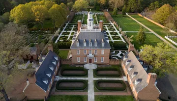 The image shows an aerial view of a large, symmetrical brick mansion with a neatly landscaped garden featuring geometric patterns and pathways.