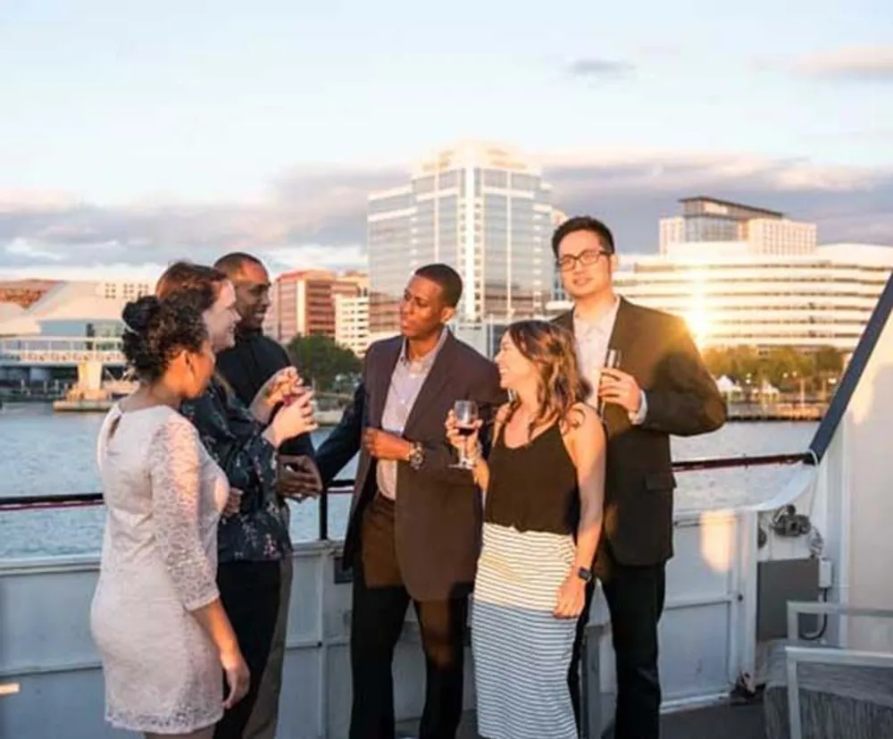 A group of people are enjoying a social gathering with drinks on a boat with a city skyline in the background during sunset