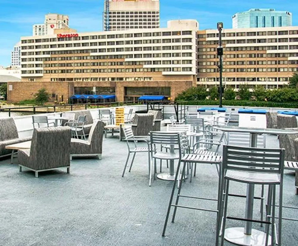 This image shows an empty outdoor seating area with modern furniture on a terrace overlooking a cityscape with buildings and a Sheraton hotel in the background