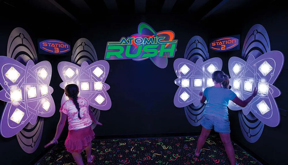 Two children are engaged in an interactive light-based game called Atomic Rush where they touch illuminated panels at different stations