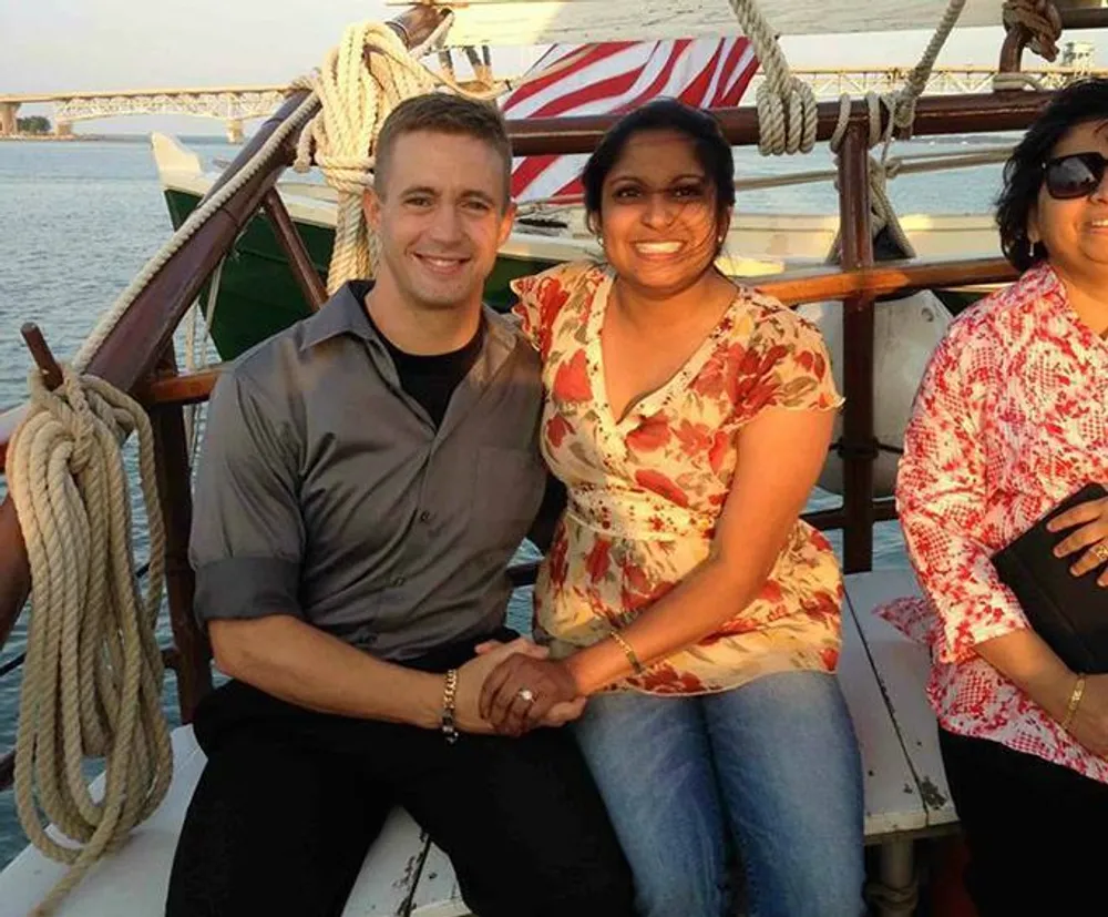 Three people are sitting on a sailboat with two of them smiling for the camera while holding hands and the American flag is visible in the background