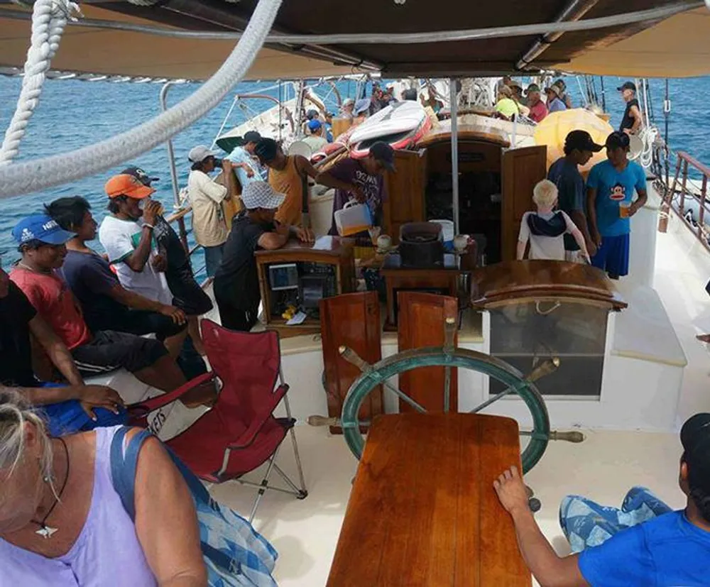 The image depicts a group of people on a boat deck with some sitting and others standing engaged in various activities under a clear sky on what appears to be a calm sea