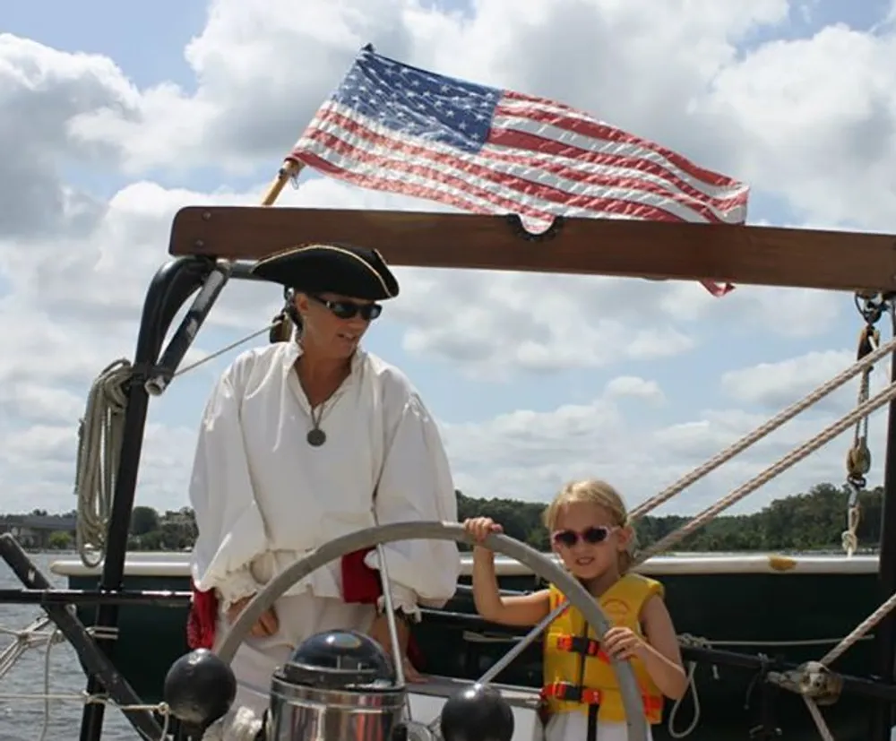 A child wearing a life jacket and sunglasses appears to be steering a boat under the guidance of an adult dressed in pirate attire with an American flag fluttering in the background