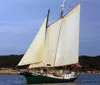 A classic sailboat with its sails unfurled is cruising on calm waters near a landmass with several people visible on board