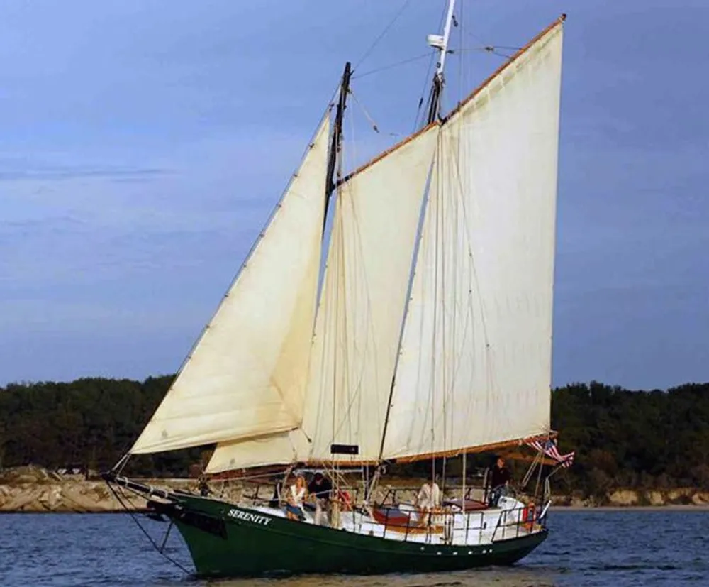A classic sailboat with its sails unfurled is cruising on calm waters near a landmass with several people visible on board