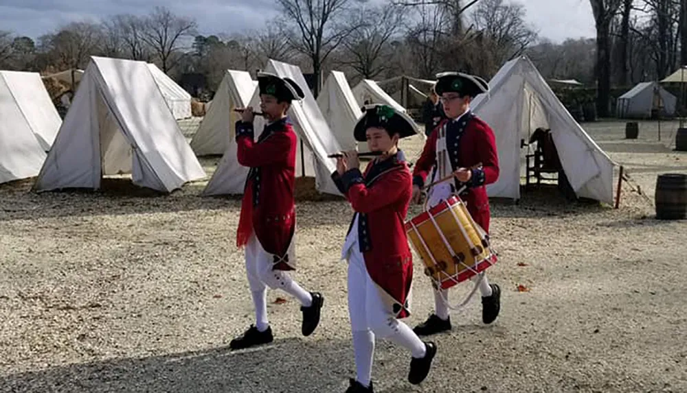Individuals dressed in historical military uniforms march with a fife and drums in a camp setting with canvas tents