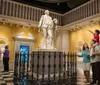 Visitors are interacting with a large statue of a historical figure in a museum-like setting with a child on someones shoulders pointing towards the sculpture