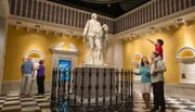 Visitors are interacting with a large statue of a historical figure in a museum-like setting, with a child on someone's shoulders pointing towards the sculpture.