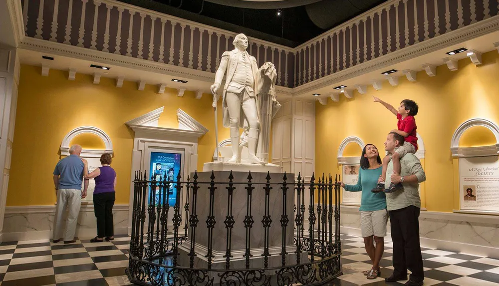 Visitors are interacting with a large statue of a historical figure in a museum-like setting with a child on someones shoulders pointing towards the sculpture