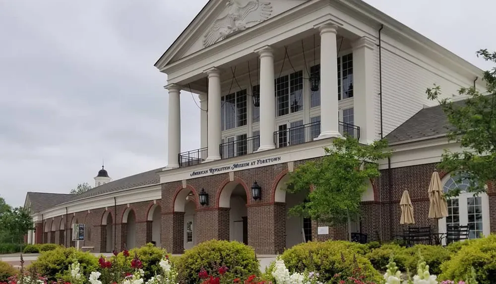 The image shows the American Revolution Museum at Yorktown featuring an elegant building with a classical facade surrounded by a manicured garden with blooming flowers