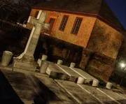 The image shows a large cross monument and several graves in a cemetery at twilight, with a historic-looking brick building in the background.