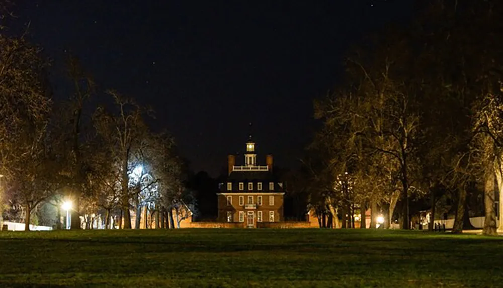 A historic brick building with a cupola is illuminated at night set against a dark sky and framed by leafless trees and a lit pathway in a grassy field