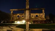 An old wooden pillory stands in the foreground of a nighttime scene, with illuminated buildings in the background under a starry sky.