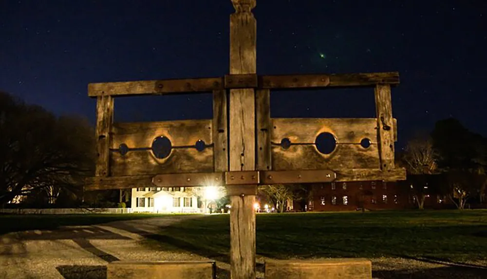 An old wooden pillory stands in the foreground of a nighttime scene with illuminated buildings in the background under a starry sky