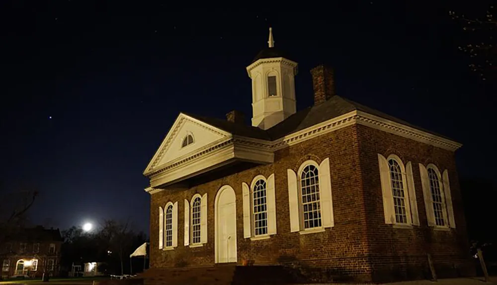 The image captures a historic brick building at night illuminated by warm lights with a clear night sky and full moon above