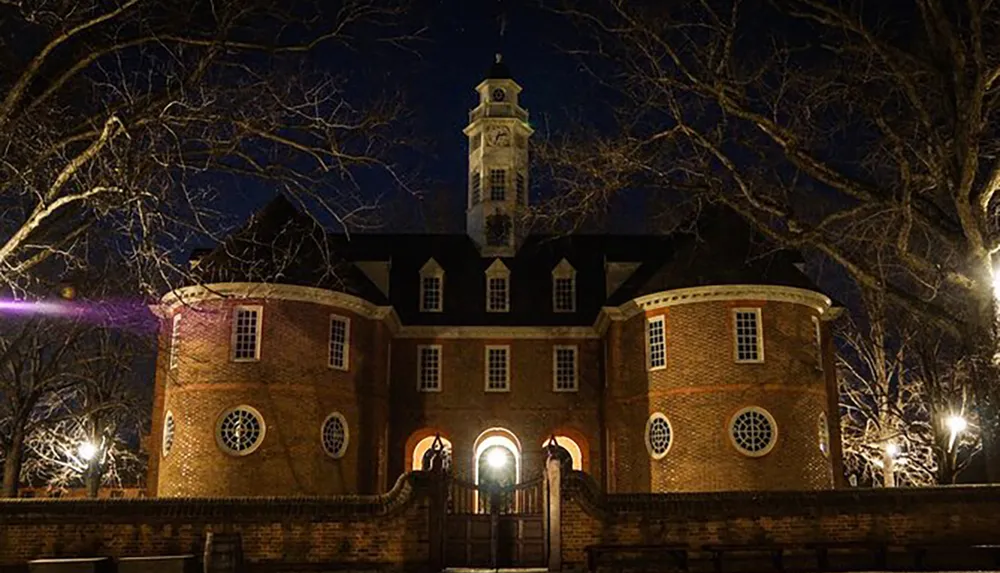 A historic brick building with a bell tower is illuminated at night casting a warm glow against the dark sky and bare tree branches