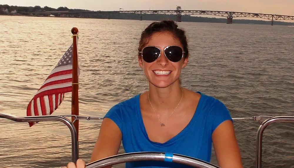 A smiling person is steering a boat with an American flag displayed in the background