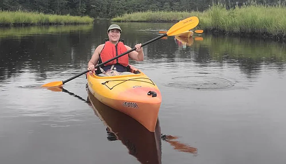 A person wearing a life vest is smiling while kayaking on calm water surrounded by greenery