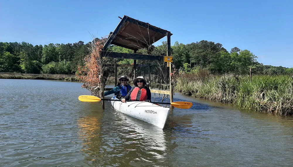 Two people are enjoying a sunny day kayaking on a calm body of water near a rustic wooden structure