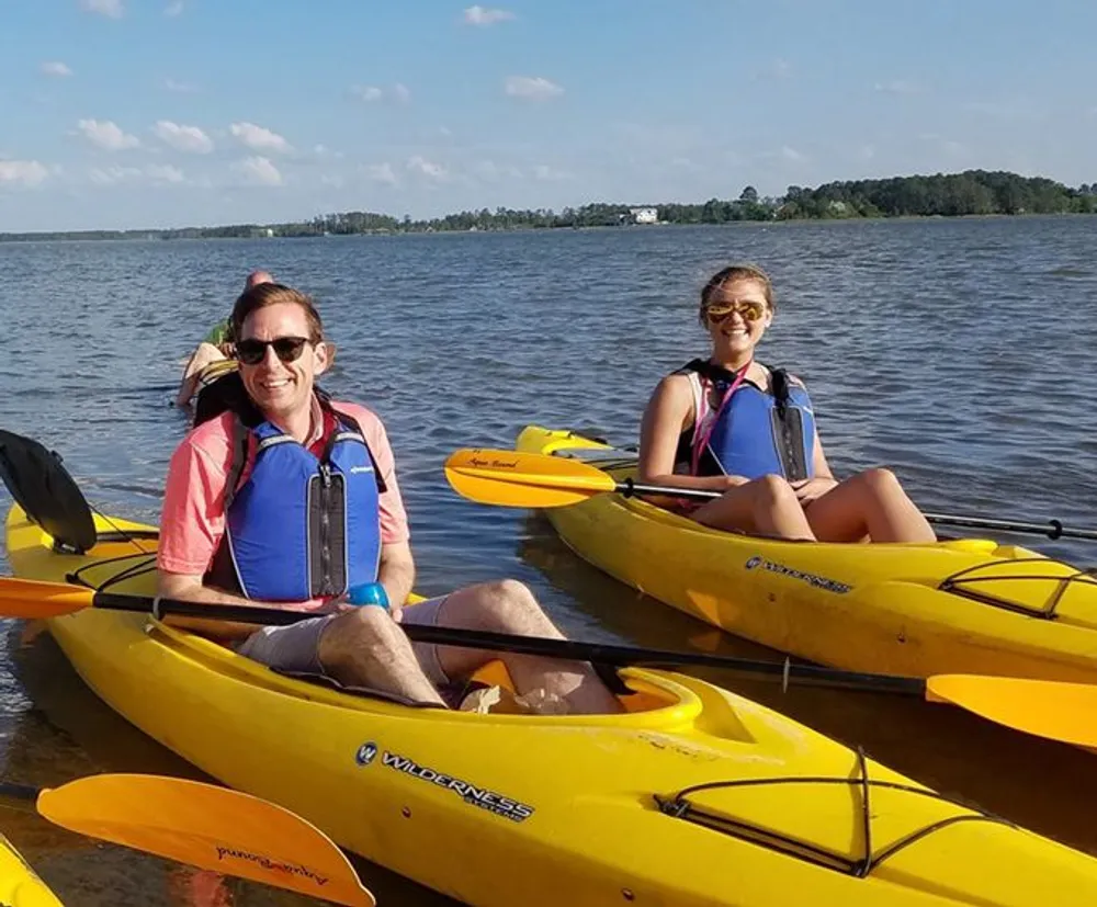 Two people are smiling while sitting in yellow kayaks on a sunny day with a body of water and trees in the background