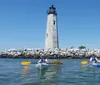 Two people in kayaks are paddling near an old lighthouse surrounded by a rocky breakwater under a clear blue sky