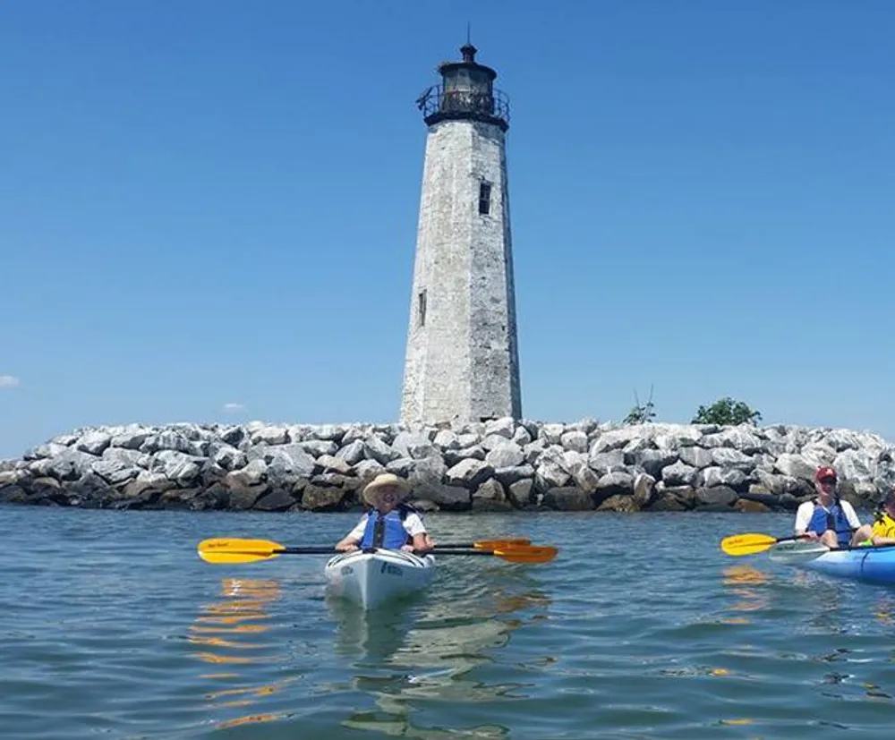 Two people in kayaks are paddling near an old lighthouse surrounded by a rocky breakwater under a clear blue sky