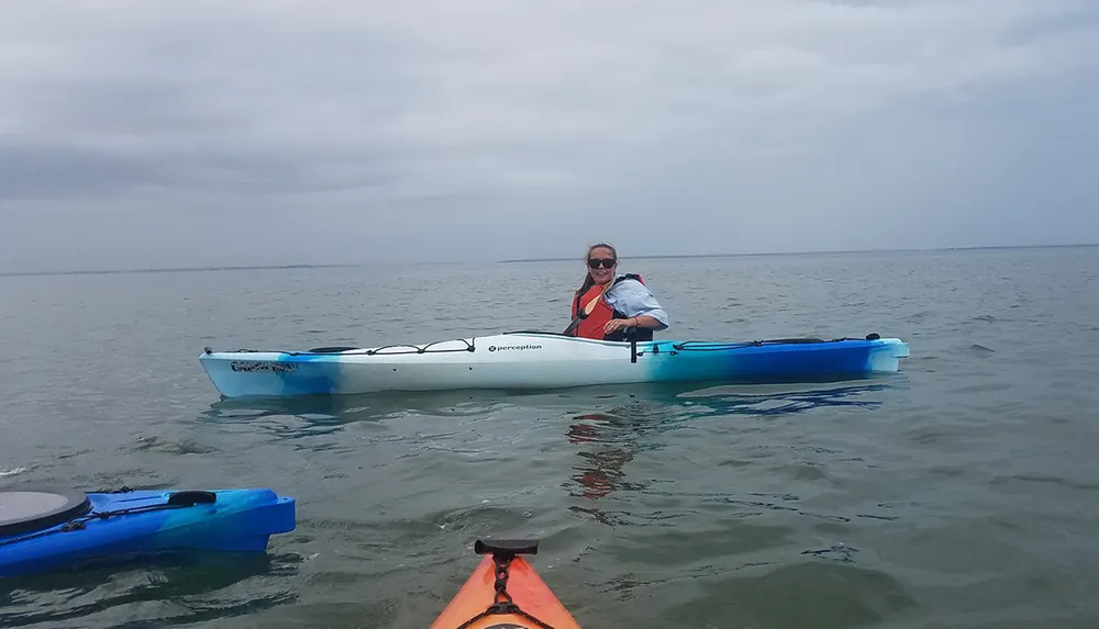 A person is smiling while kayaking on calm waters with a glimpse of another kayak in the foreground