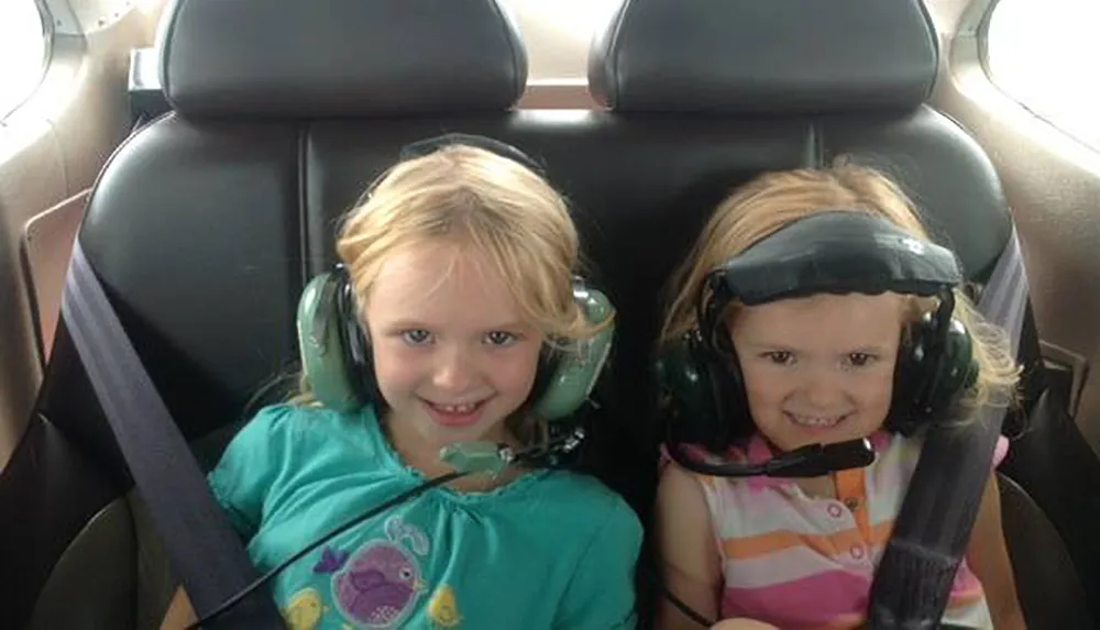 Two young children are smiling while wearing headsets in the backseat of a vehicle