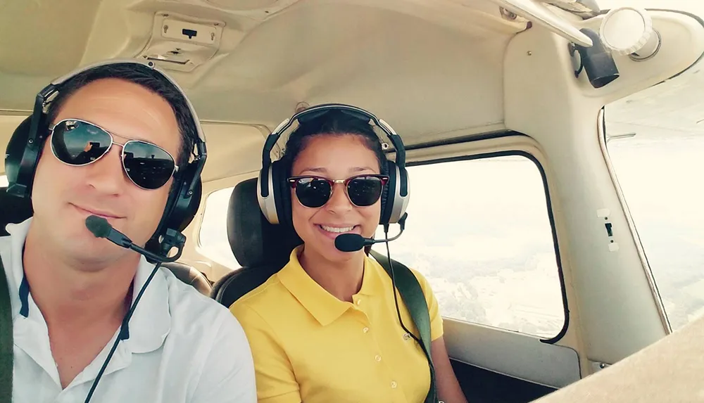 Two smiling people are wearing aviation headsets inside a small airplane with one of them presumably piloting the aircraft