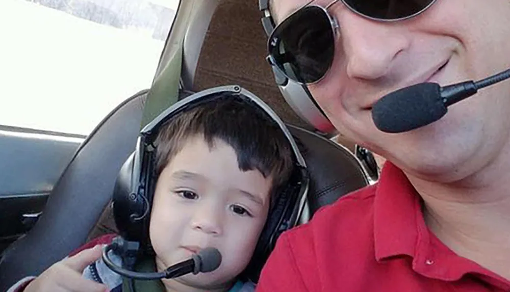 A man and a young child wearing aviation headsets are seated inside an aircraft presumably enjoying a shared experience of flying