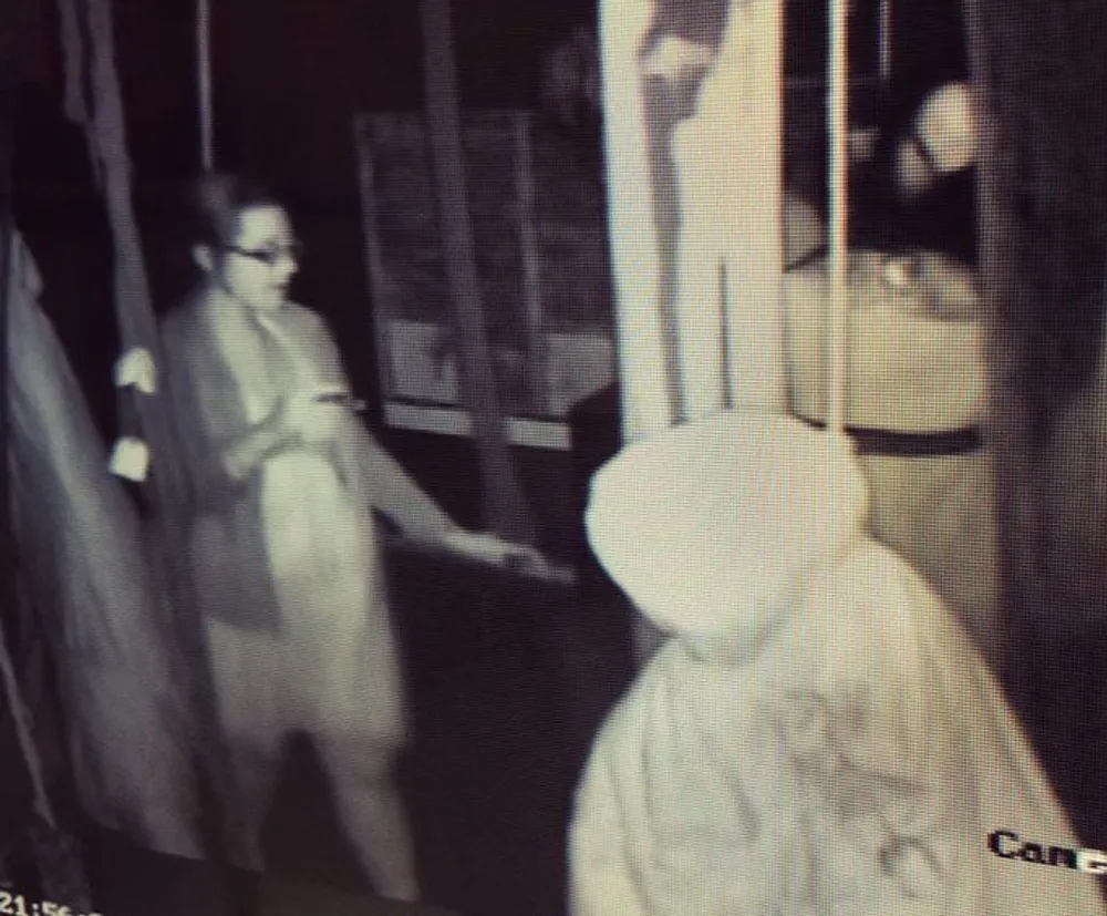 The image shows a grainy monochrome CCTV footage of a person in a hooded garment shaking hands with another person wearing glasses seemingly indoors