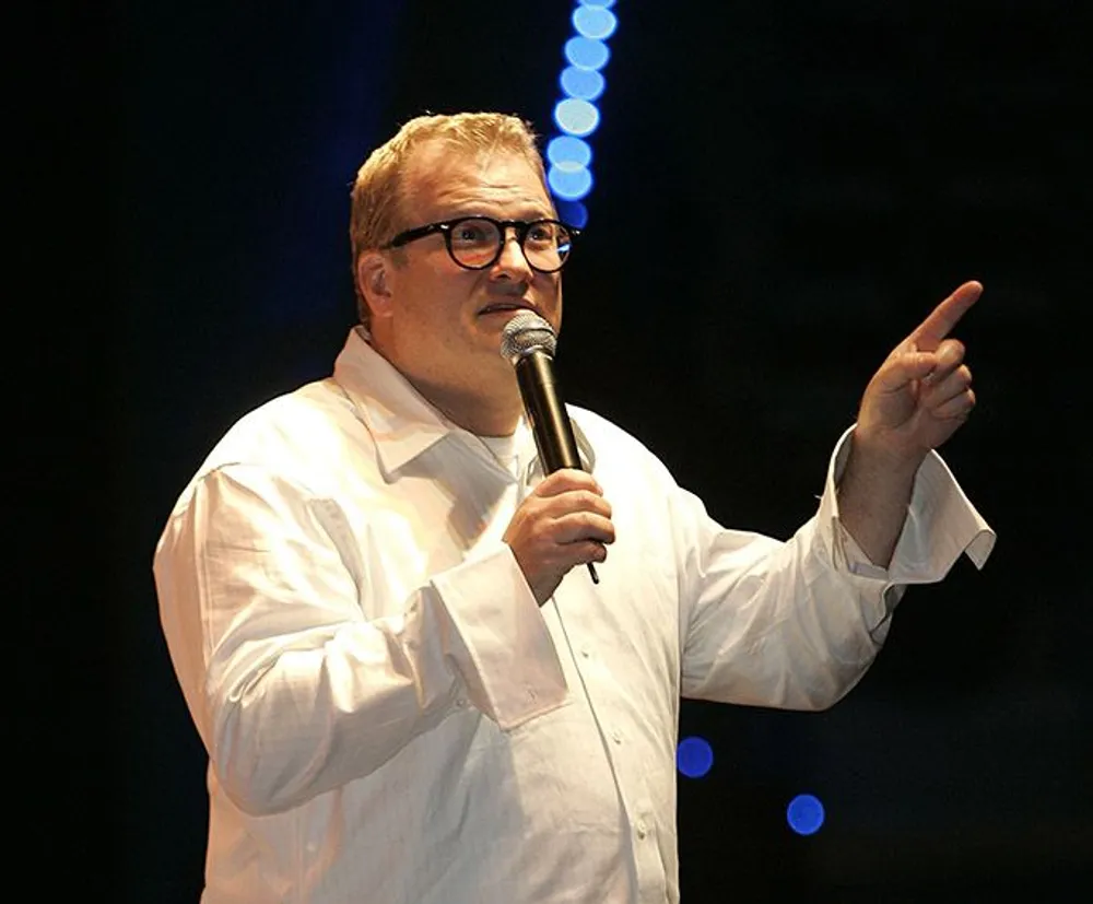 A man in glasses wearing a white shirt is speaking into a microphone and pointing to the side while standing on a stage with blue lights in the background