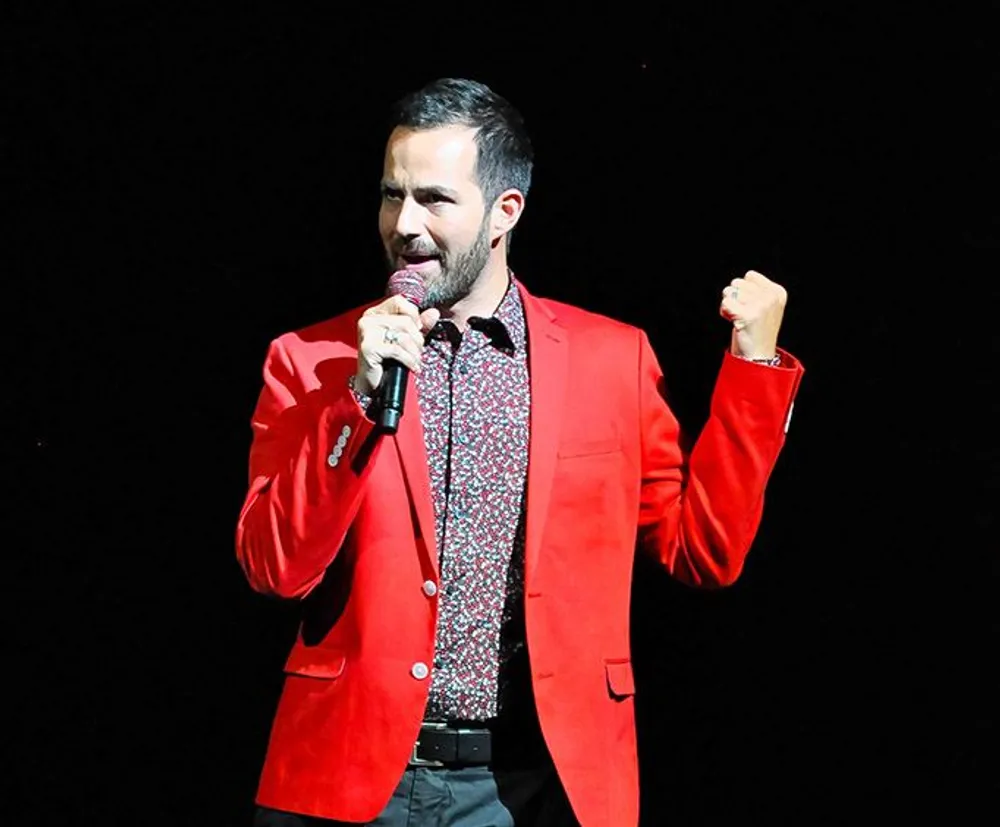 A person is performing on stage wearing a red jacket and holding a microphone with a fist raised in what appears to be a moment of expression or emphasis