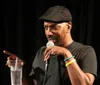 A person in a black cap is speaking into a microphone with a smile gesturing with one hand while holding a glass of water in the other