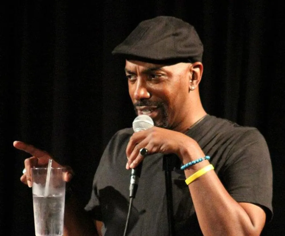 A person in a black cap is speaking into a microphone with a smile gesturing with one hand while holding a glass of water in the other
