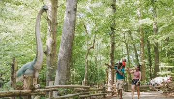 A family is interacting with a life-size dinosaur model in a forested outdoor setting.