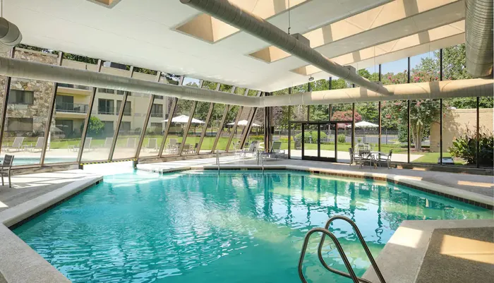 The image shows an indoor swimming pool with clear blue water flanked by loungers and large windows that provide a view of a sunny outdoor setting